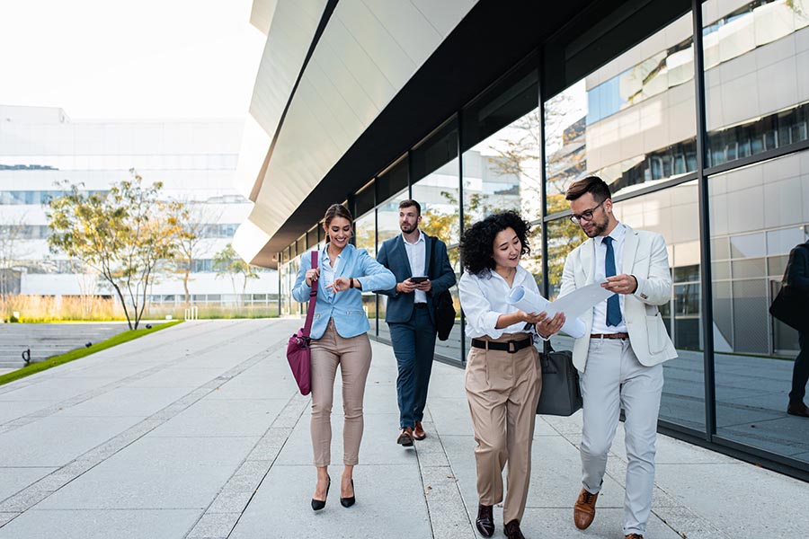 Contact Us - Four Colleagues Stroll Alongside Their Office Building, Reviewing Plans and Checking the Time